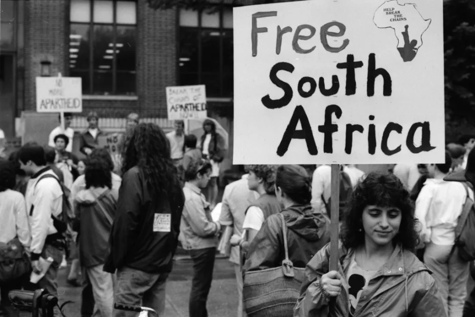 黑白照片:一名年轻女子举着写有「Free South Africa」和非洲版图图案的标语牌,背景人群中还有人举着「No More Apartheid」等标语,众人聚集在一栋建筑前。