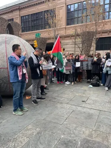 户外广场上聚集着手持标语牌的人群,一面巴勒斯坦国旗竖立其中,前方两名男子面对镜头打着手语,广场上有一座巨大的球形雕塑,背景是欧式风格建筑。