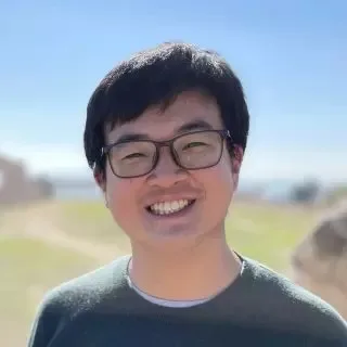 一名戴眼镜、穿深绿色上衣的男子微笑面对镜头,背景是户外虚化的草地与天空。
