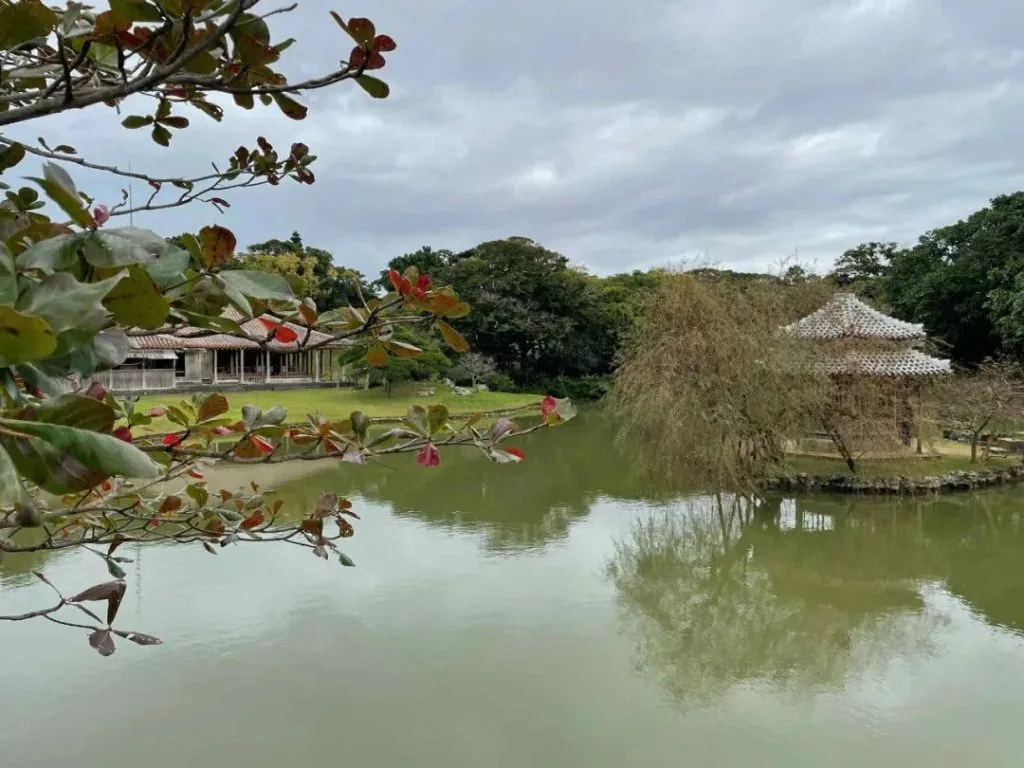 一池碧水环绕,对岸是茅顶六角亭和一座低矮瓦顶建筑,池边垂柳依依,近处枝头缀着红叶。