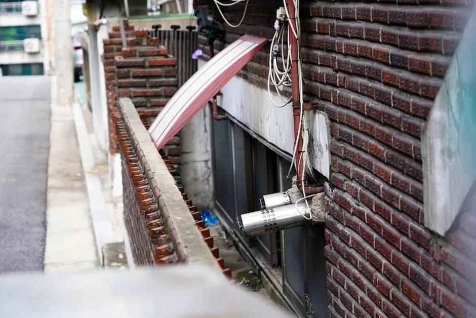 红砖建筑沿坡道而建,外墙缠绕着电线和管道,靠近地面处是带金属护栏的半地下室窗户,窗下堆着杂物。