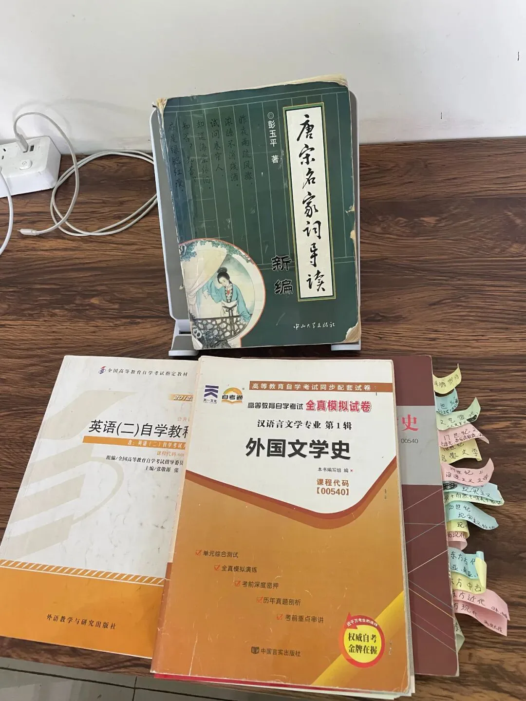 书桌上摆放自考备考书籍的照片,前排竖立《唐宋名家词导读新编》,其后是《英语(二)自学教程》和贴满彩色标签纸的《外国文学史》全真模拟试卷。