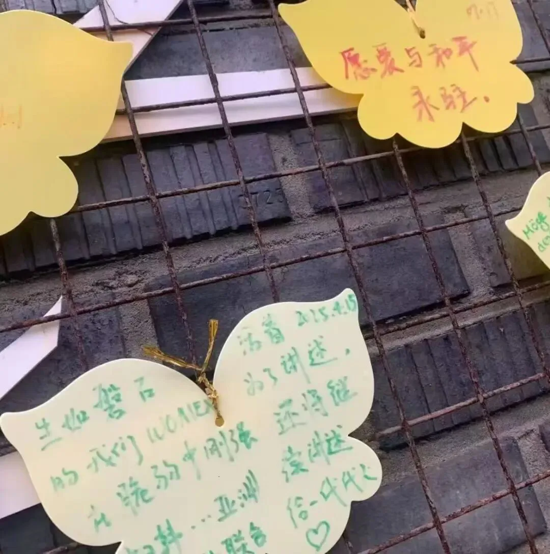蝴蝶形彩色纸片系在锈迹斑斑的金属网格上,纸片上手写留言,其中一张写着“愿爱与和平永驻”,另一张提到“WOMEN”及持续讲述诉求,其余字迹潦草不易辨认。