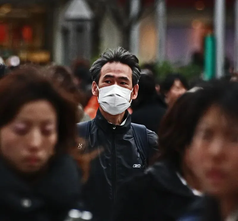 拥挤人群中，一名戴白色口罩的男子清晰对焦，周围行人面部模糊。