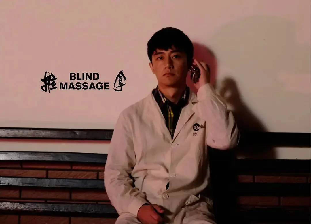 剧照中一名男子坐在木椅上,身穿米色外套,手持手机贴在耳边,目光望向前方;画面左侧印有片名《推拿》及英文 BLIND MASSAGE。