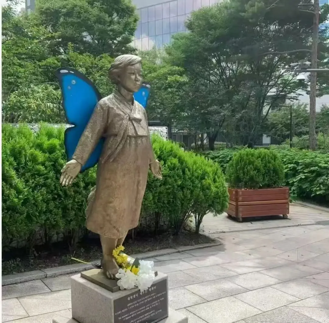 青铜色雕像身着传统韩服站立,身后展开一对蓝色蝴蝶翅膀,脚边摆放黄白色鲜花,四周环绕修剪整齐的绿植和石板广场。