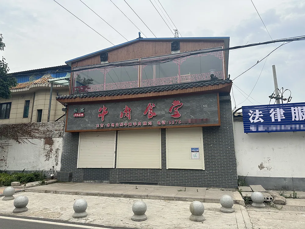 一栋两层小楼,招牌写着“牛肉食堂”,卷帘门紧闭,门前摆着石球路障,旁边是一块“法律服务”蓝色招牌。