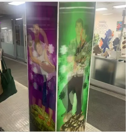 两块紫色和绿色调的透镜印刷展板立在瓷砖地面的通道中,紫色一面印着怀抱婴儿的女性,绿色一面印着站立的年轻男子,背景衬着绿叶图案,通道尽头摆着毛绒玩偶等小物件。